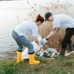 Two volunteers pick up trash by a river, promoting environmental conservation.